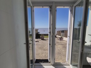 A view through a glass door to a patio with a view of the sea, potted plants, and a concrete ground. - Beautiful oceanfront apartment - sleeps 3 (FOURAS)