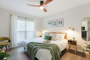 Un dormitorio con cama, ventilador de techo, mesita de noche con lámpara, espejo y ventana con cortinas.