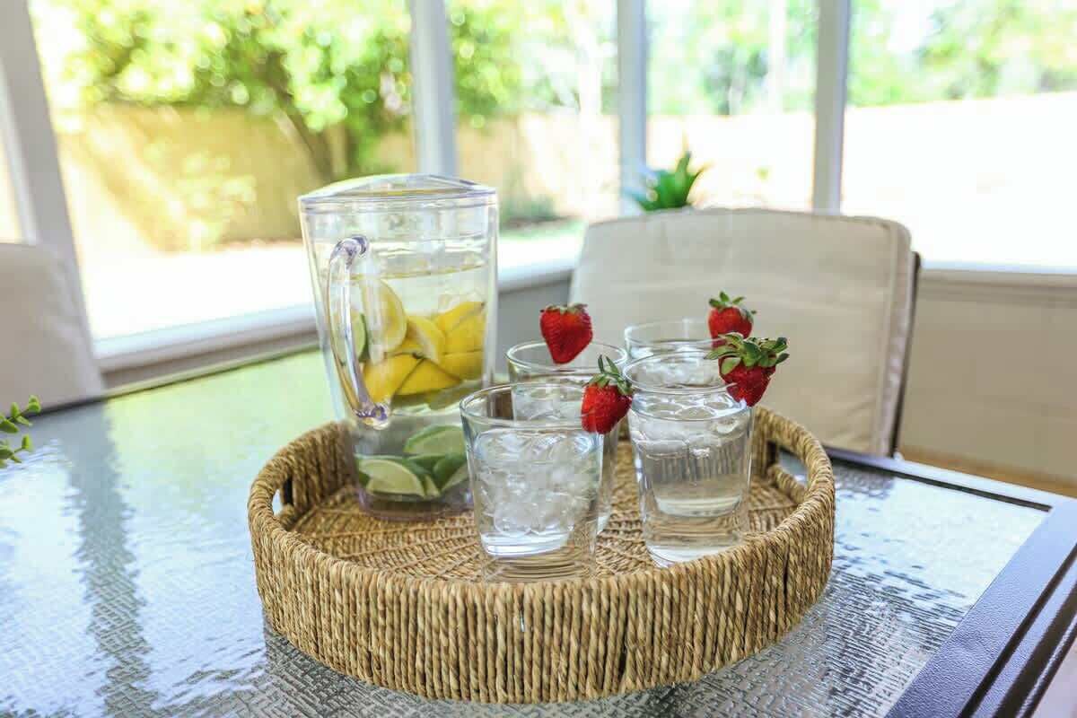 A glass pitcher with lemon slices and ice, two glasses with strawberries, and a woven tray on a table by a window.