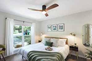 Un dormitorio con cama, ventilador de techo, ventana con cortinas, mesita de noche con lámpara y espejo.