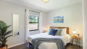 Un dormitorio con cama, mesitas de noche, ventana, espejo, planta y un cuadro en la pared.