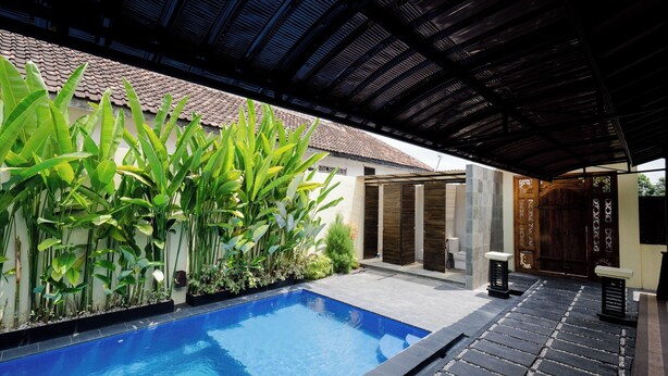 Una piscina con solárium de baldosas, rodeada de exuberante vegetación y una zona cubierta con techo de baldosas.