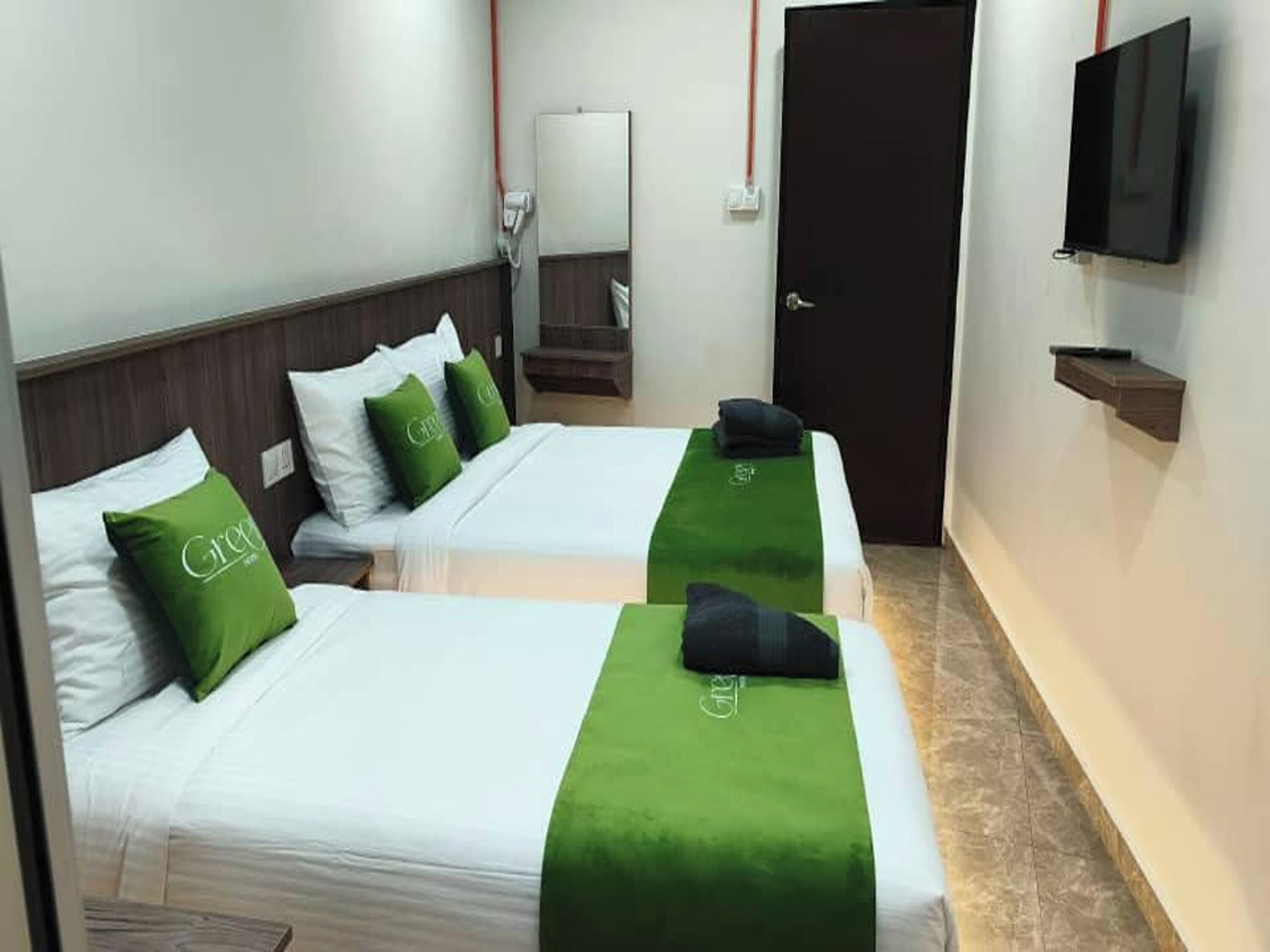 A hotel room with two beds, each with a green bedspread and matching pillows, a flat-screen TV mounted on the wall, and a door with a red handle.