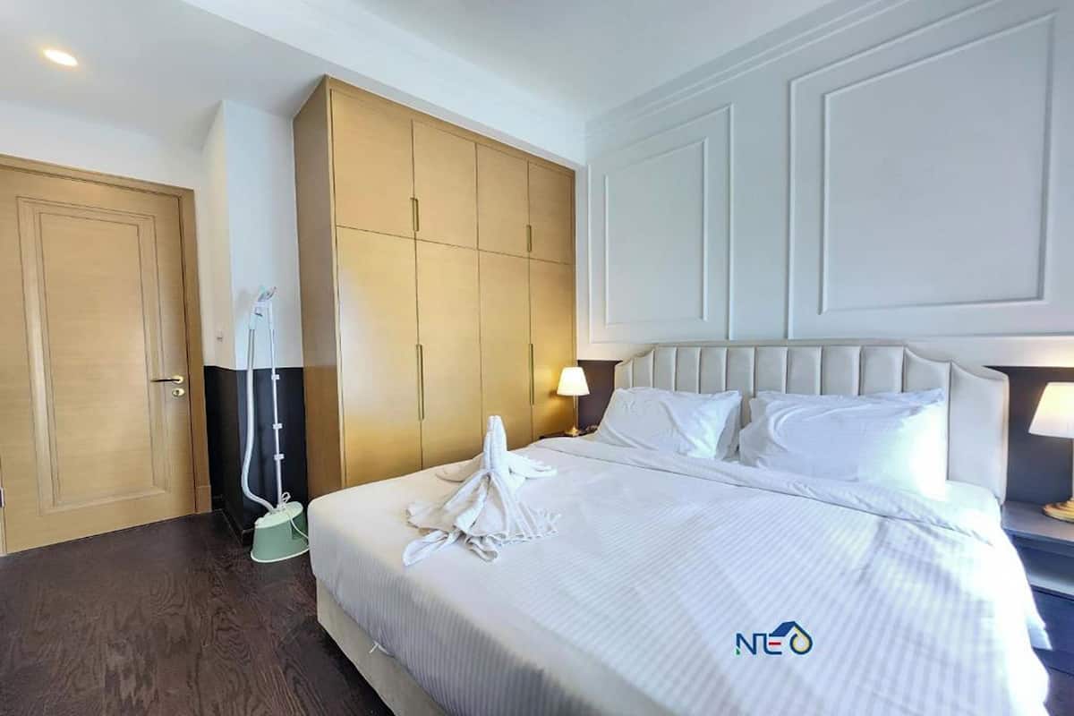 A hotel room with a large bed, a wardrobe, a vacuum cleaner, and a nightstand with a lamp.
