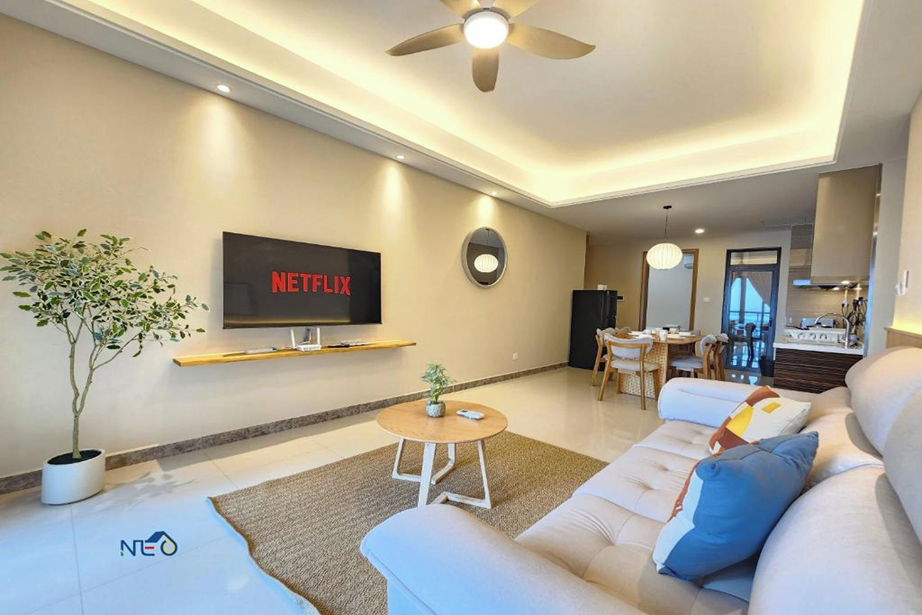 A modern living room with a flat-screen TV, a ceiling fan, a dining area, and a kitchen in the background.