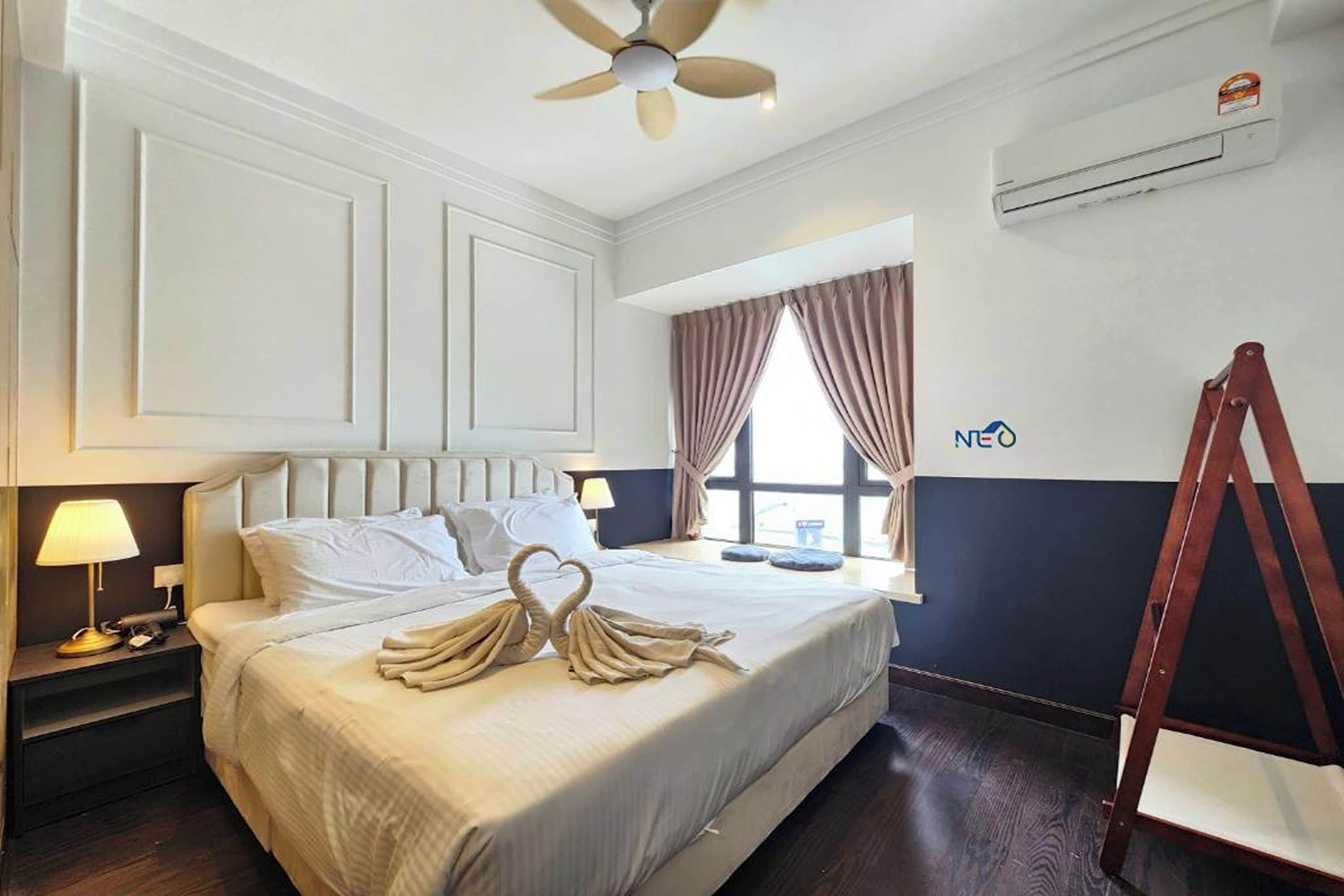 A hotel room with a bed, a ceiling fan, an air conditioning unit, a window with curtains, and a wooden chair.