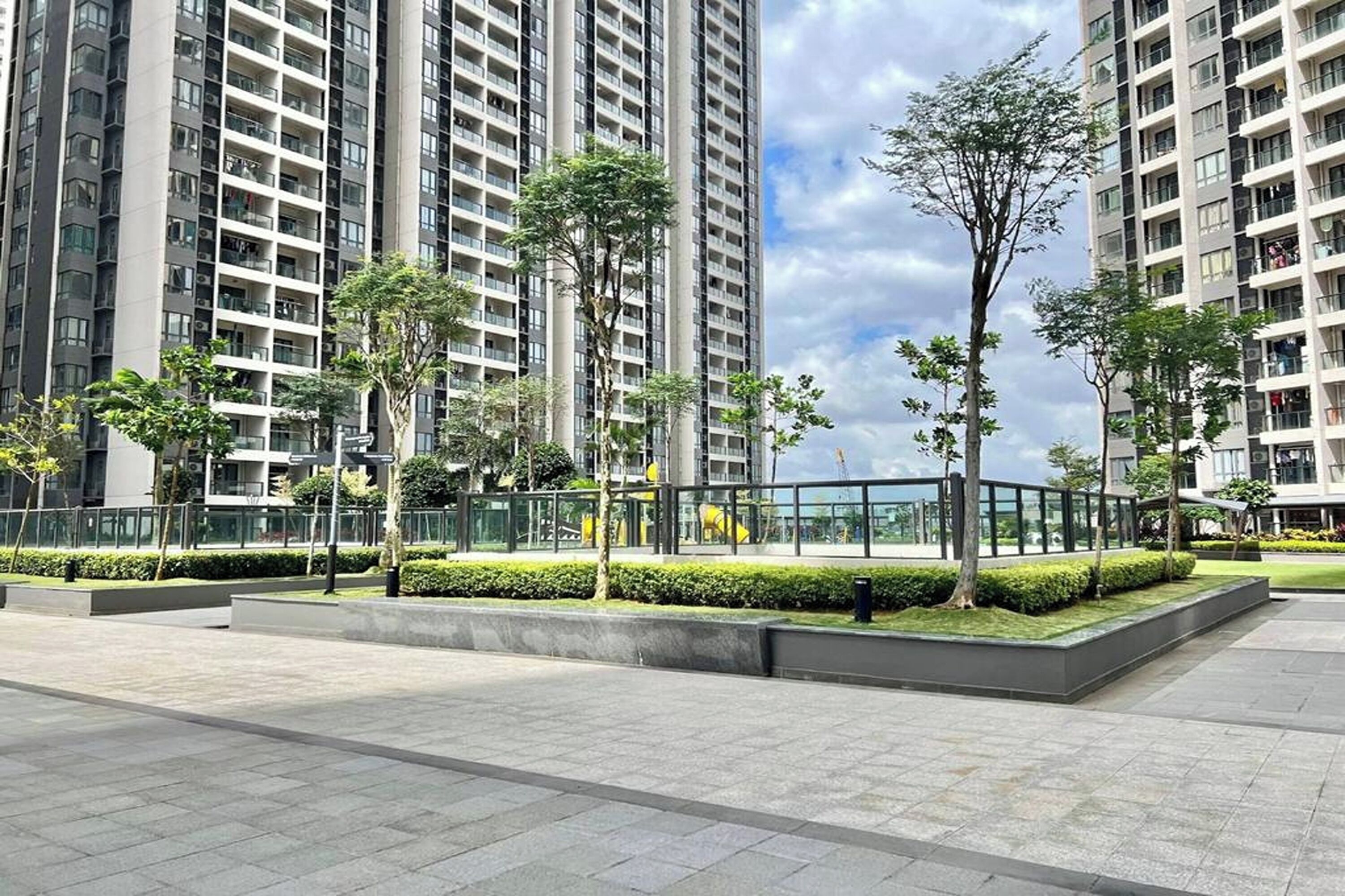 A modern urban residential area with high-rise buildings, landscaped greenery, and a paved walkway.