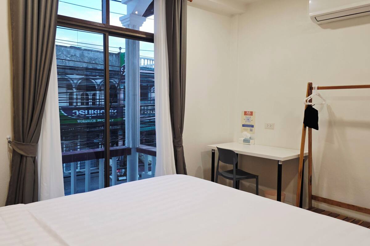 Quarto de hotel com cama, escrivaninha, cadeira, janela com cortinas e vista para a paisagem urbana.