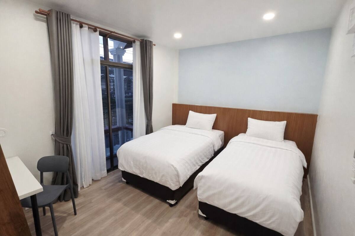 Quarto de hotel com duas camas, uma escrivaninha, uma cadeira e uma janela com cortinas.