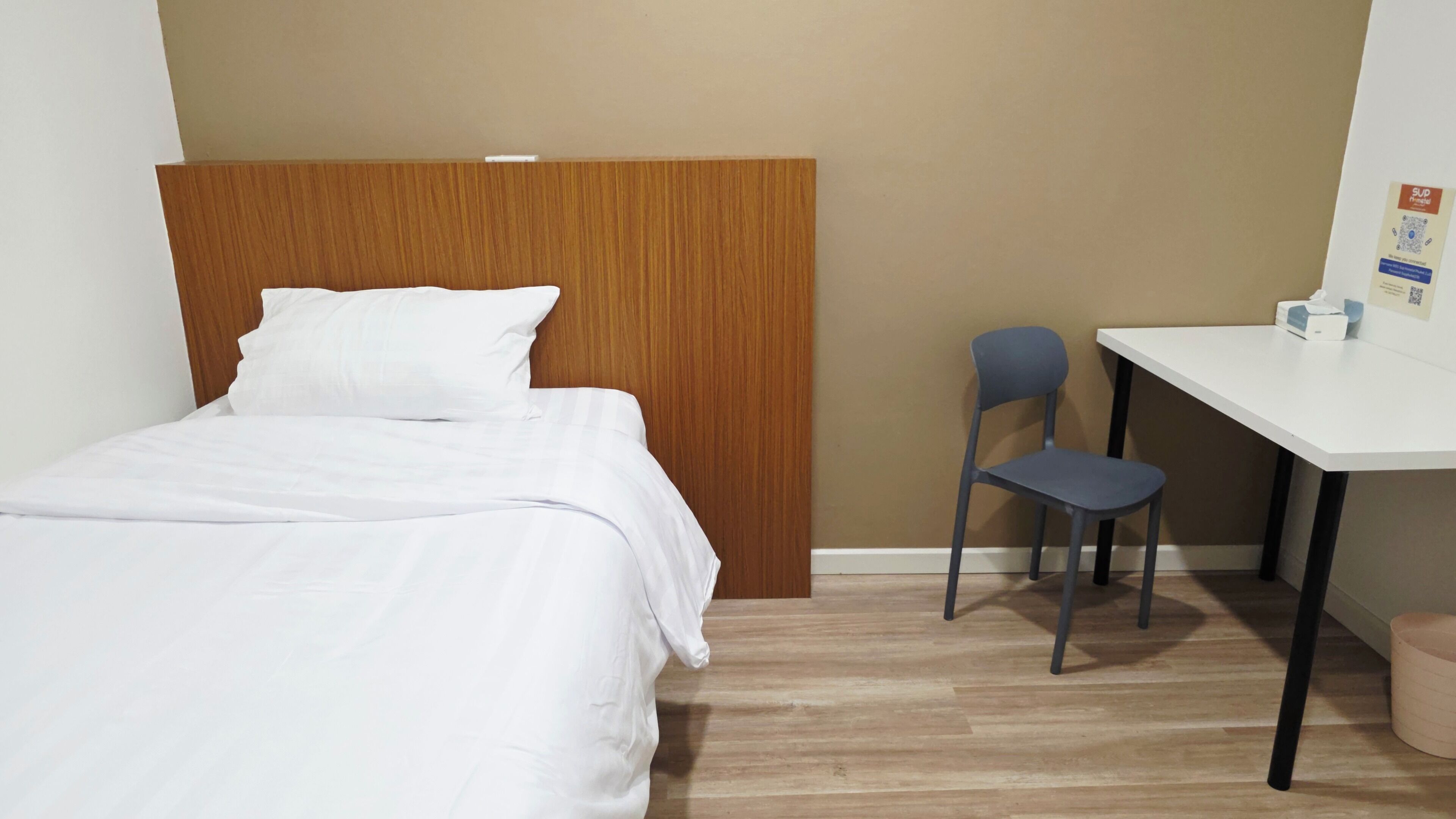 A hotel room with a bed, a wooden headboard, a desk, a chair, and a small trash bin.