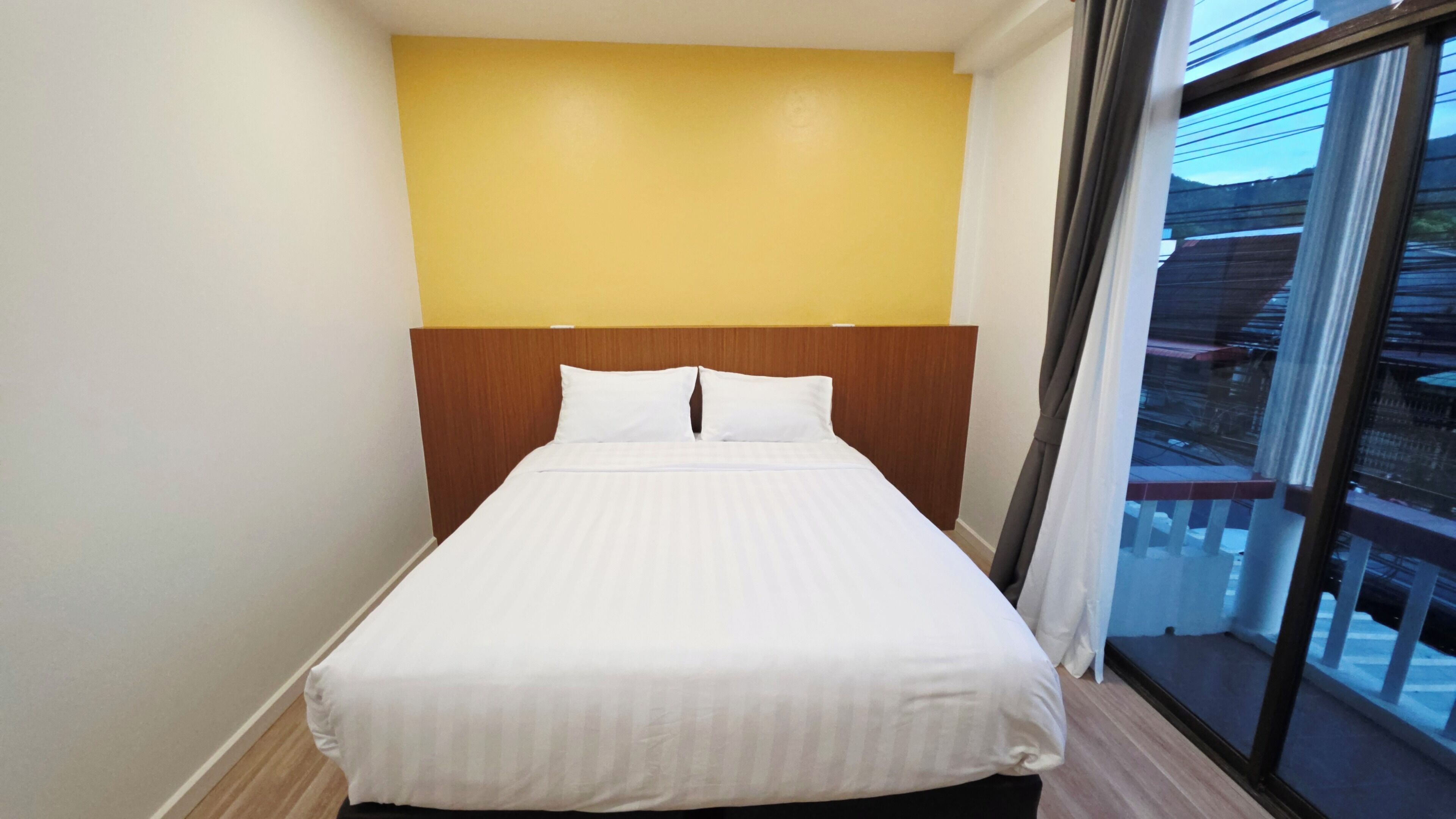 Een eenpersoonsbed in een kamer met een houten hoofdbord, een gele wand en een raam met uitzicht op gebouwen.