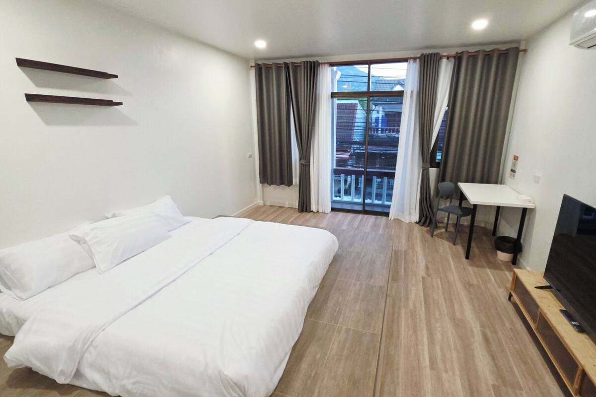 Quarto de hotel com cama, televisão, escrivaninha e varanda com cortinas.
