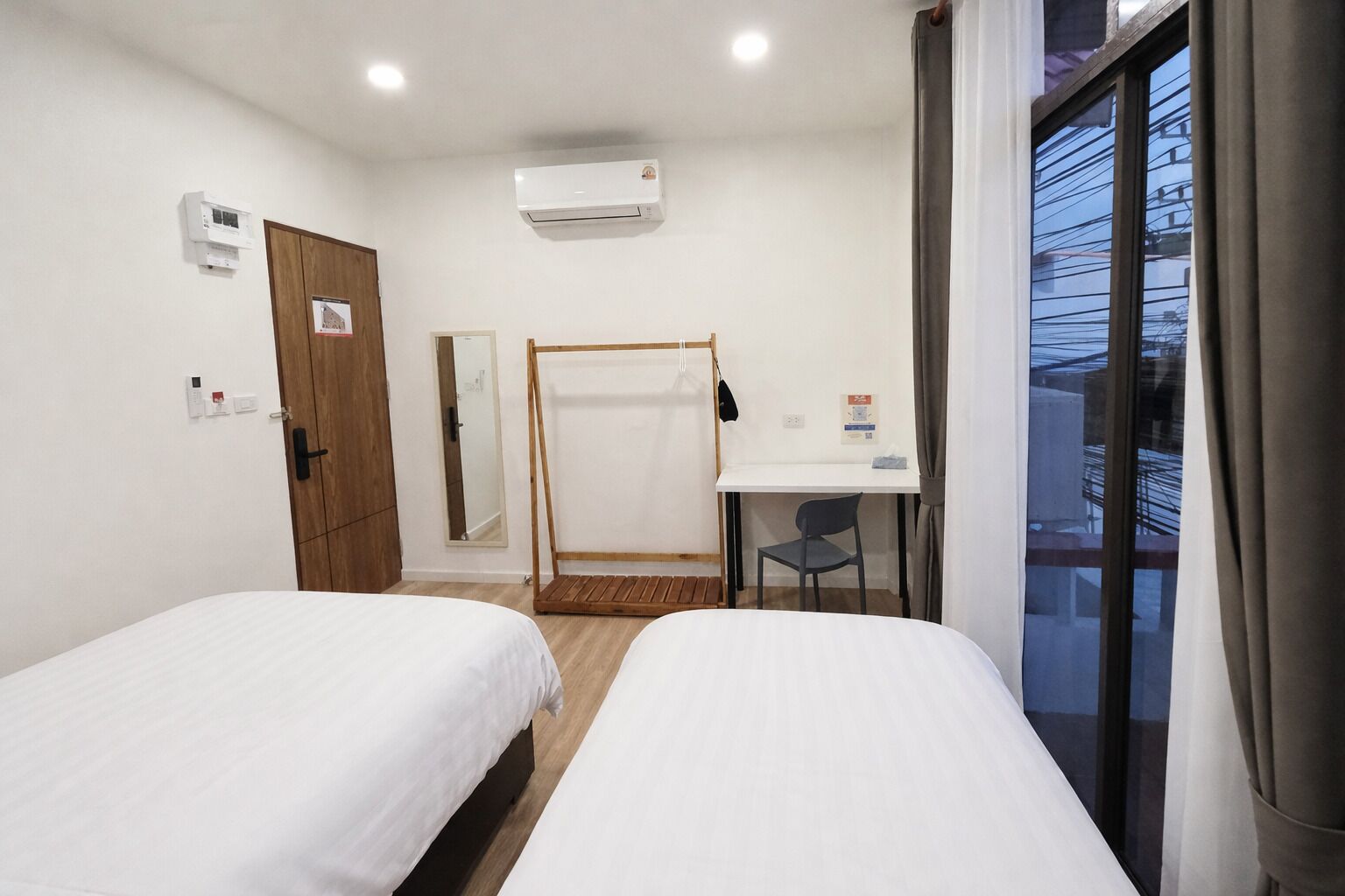 Une chambre d’hôtel avec deux lits, un bureau, une chaise, un appareil de climatisation, une armoire en bois, un miroir et une fenêtre donnant sur l’extérieur.
