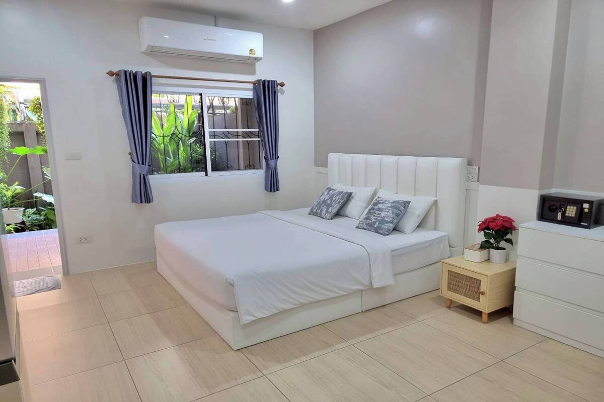 Un dormitorio con cama, una mesita de noche con planta, una ventana con cortinas y un equipo de aire acondicionado.