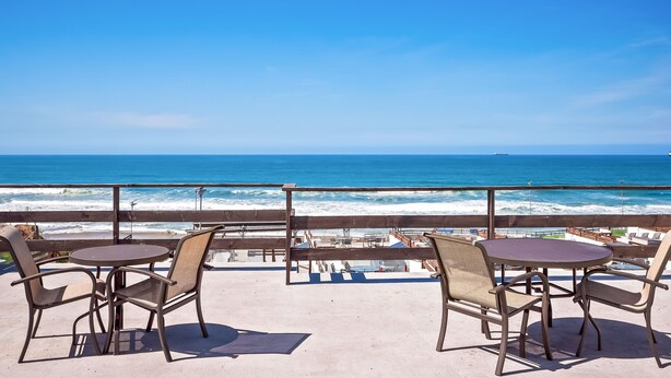 Een balkon met twee ronde tafels en vier stoelen, met uitzicht op een strand met golven en een helderblauwe lucht.