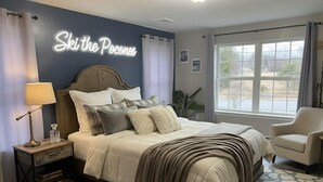 Un dormitorio con cama, mesitas de noche, lámpara, silla, ventana y un cartel en la pared.