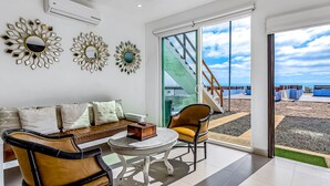Ein modernes Wohnzimmer mit einer Couch, zwei Sesseln, einem Couchtisch und dekorativen Spiegeln. Eine große Schiebeglas-Tür führt zu einem Balkon mit Blick auf Strand und Meer.