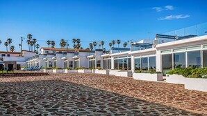 Un hotel moderno con un patio empedrado, palmeras y un cielo azul despejado.
