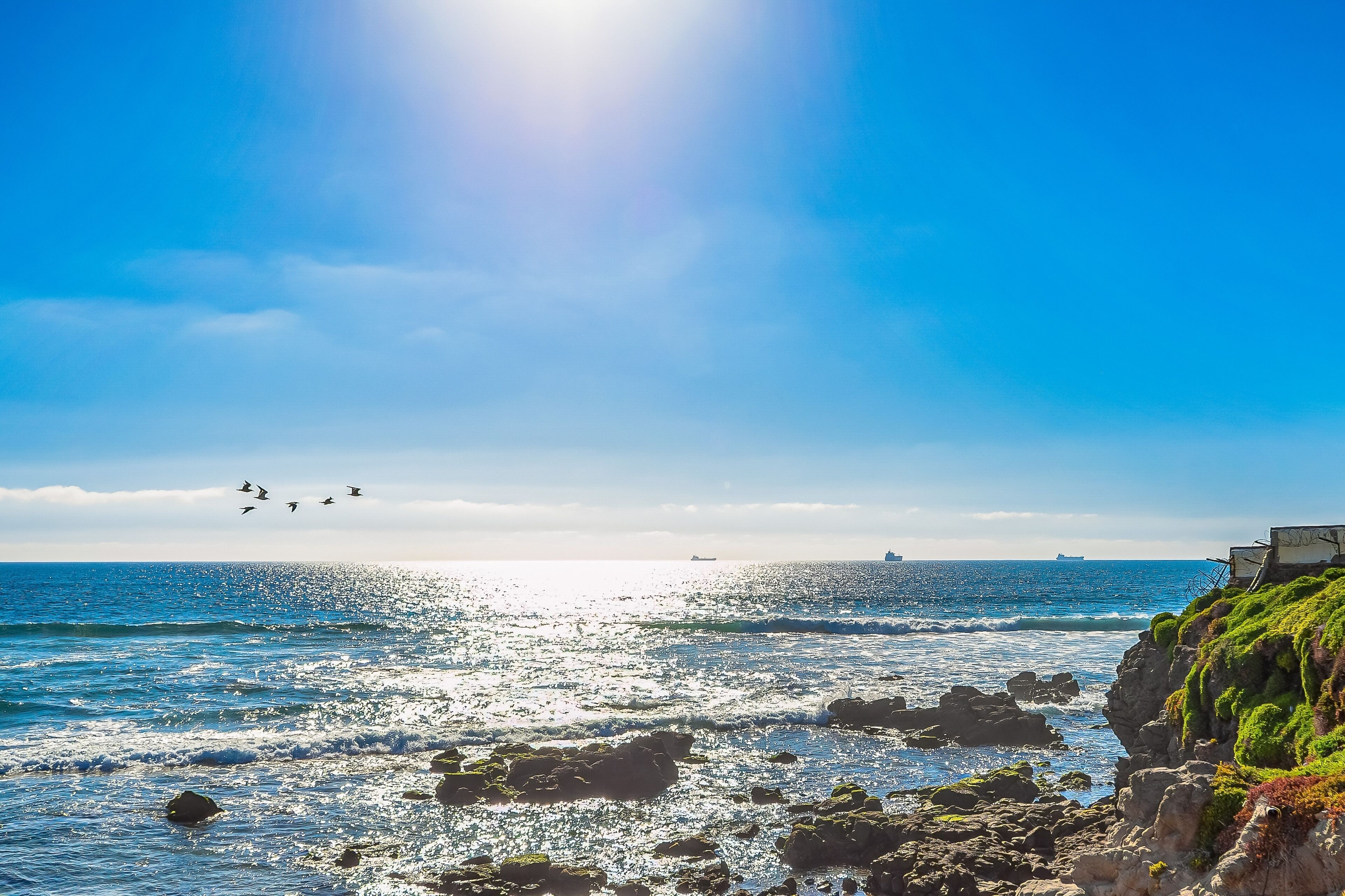 Un paysage côtier avec des rivages rocheux, un ciel bleu clair et des navires à l’horizon.