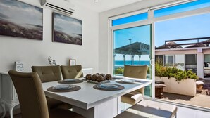 A dining area with a white table, brown chairs, and a view of the ocean through large windows. - Peaceful dog-friendly villa with outdoor pool, rooftop patio, beach views, & AC (Rosarito)