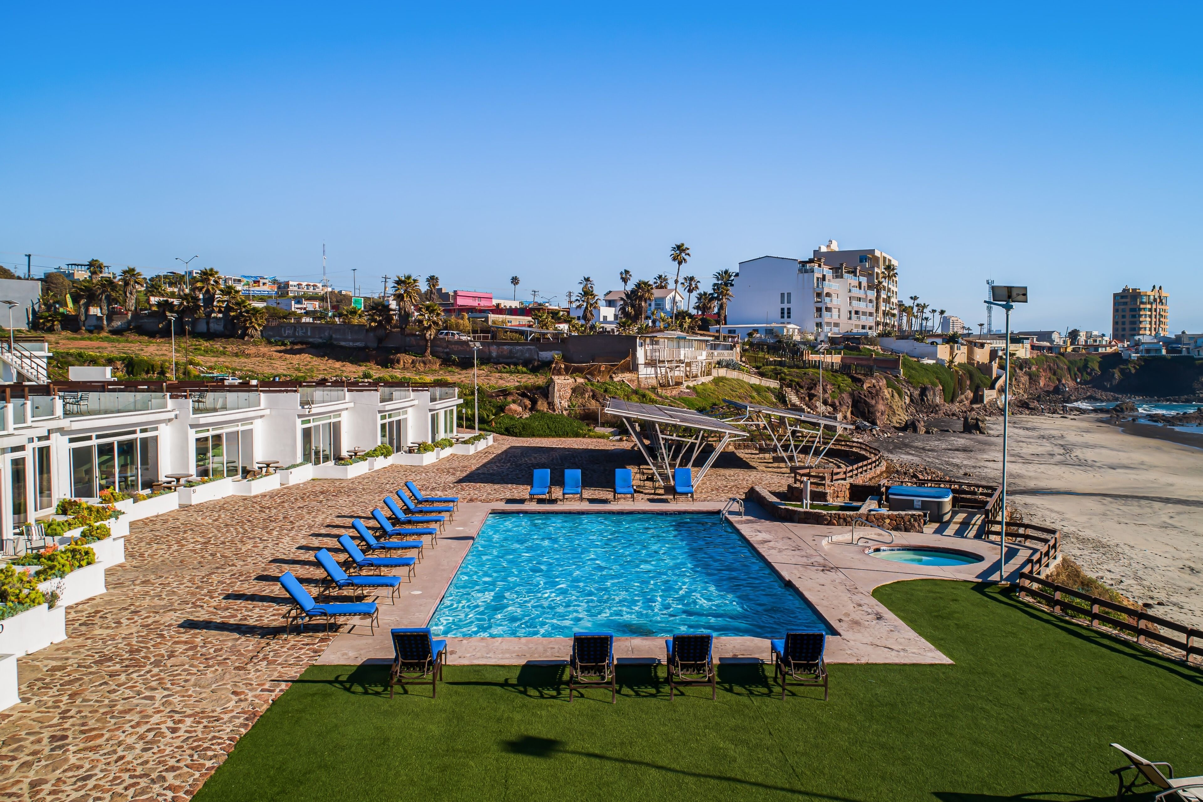 Un espace piscine agrémenté de chaises longues bleues et d’un petit jacuzzi, entouré d’un jardin aménagé et offrant une vue sur la plage et l’océan.