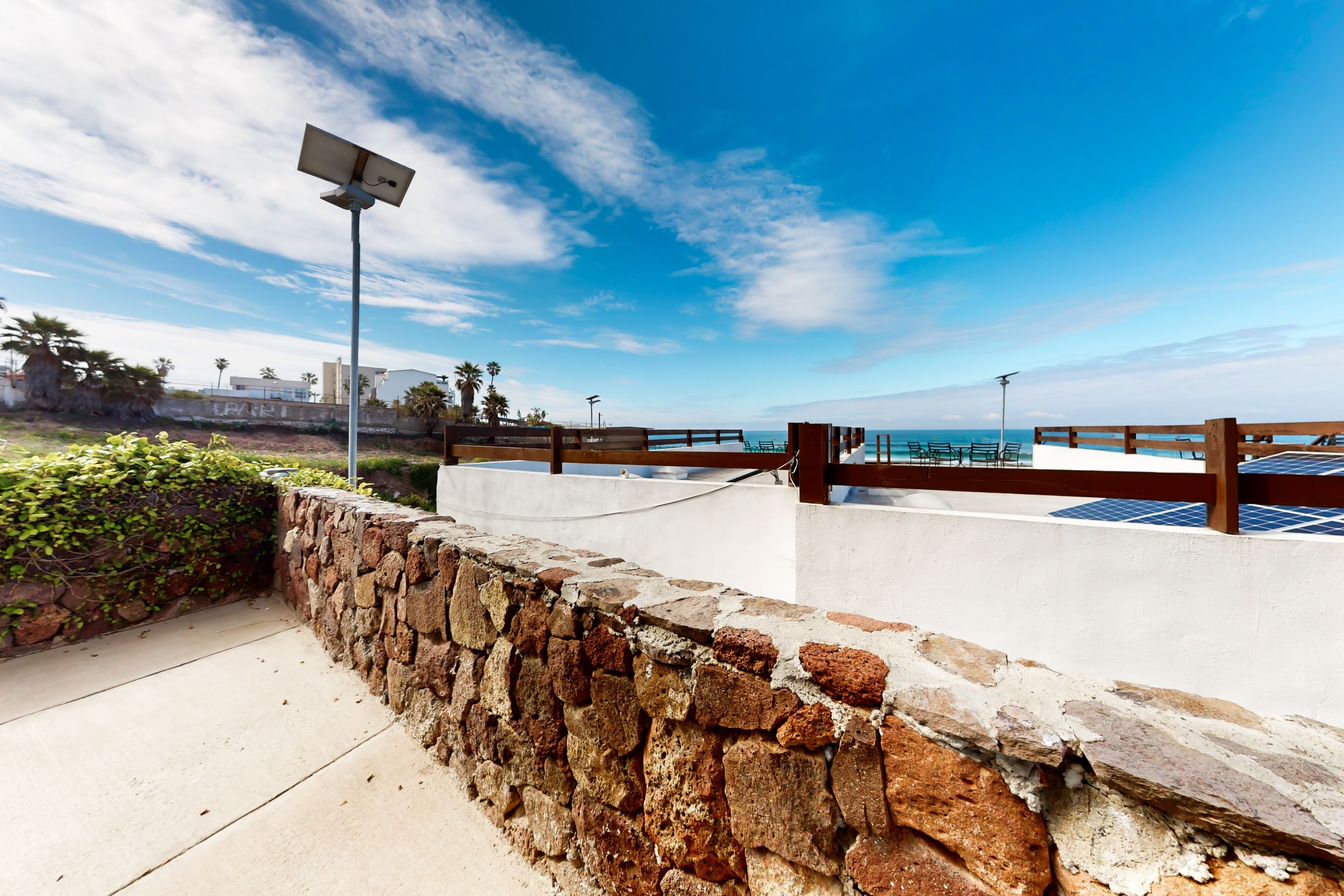 A coastal walkway with a stone wall, a wooden fence, and solar panels on a building.