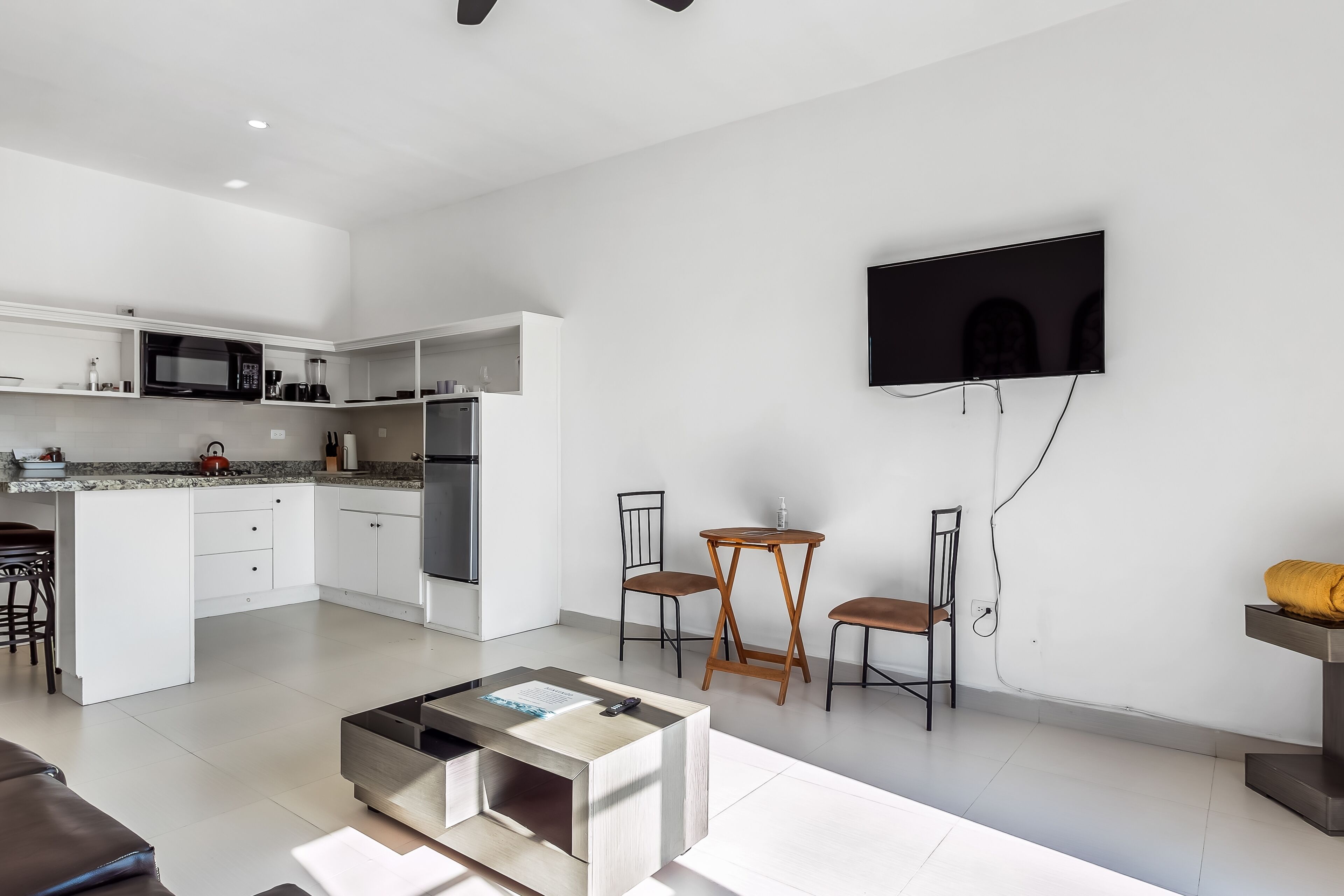Una cocina moderna con gabinetes blancos, un microondas integrado y una cafetera. Hay una zona de comedor con una mesa redonda pequeña y dos sillas. Un televisor de pantalla plana está montado en la pared.