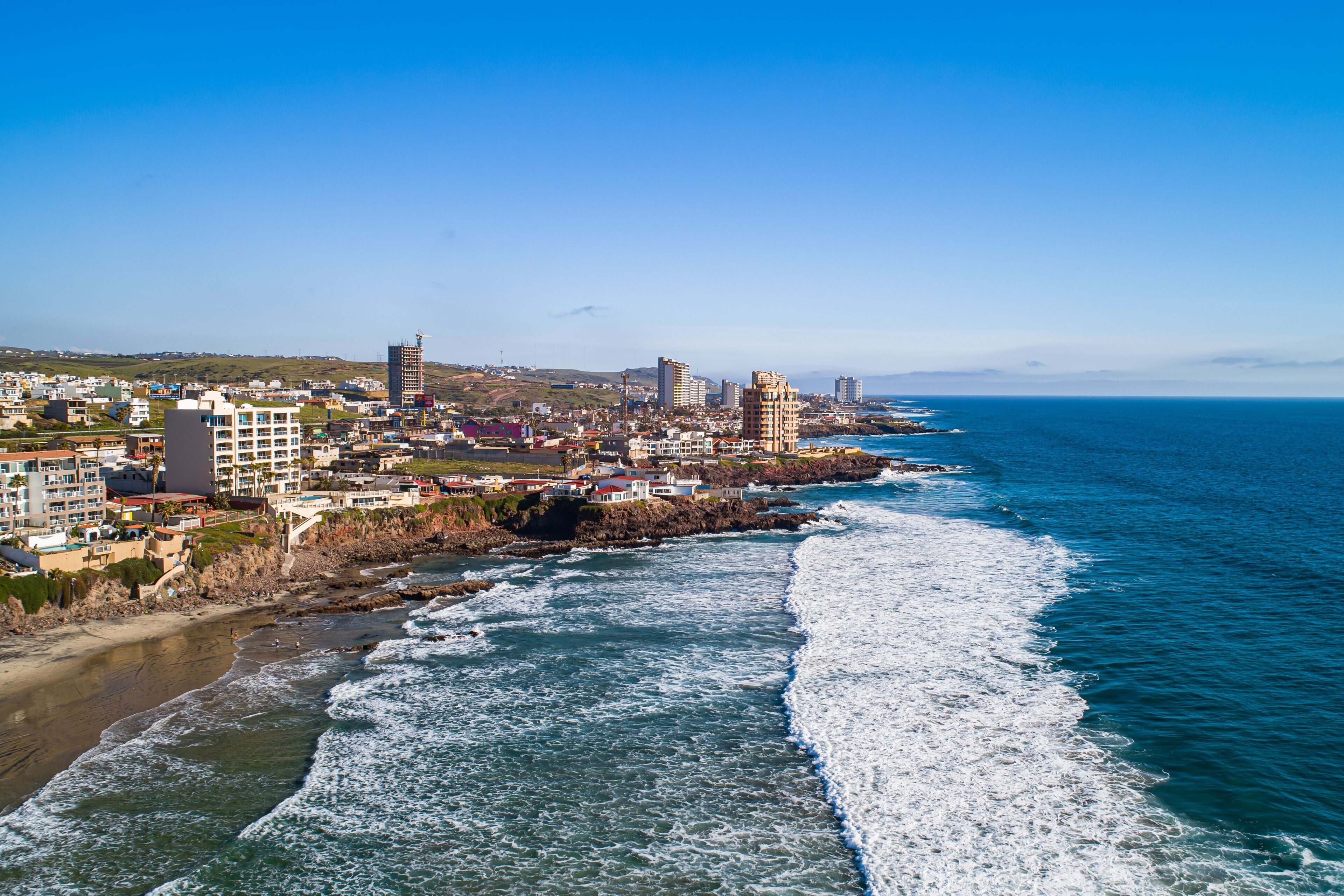 Un paysage urbain côtier avec des bâtiments le long de la côte, un ciel bleu dégagé et des vagues qui déferlent sur la plage.