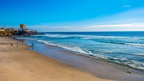 Una playa con un edificio en un acantilado, olas y cielo despejado.