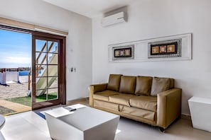 Ein modernes Wohnzimmer mit einem beigen Ledersofa, einem weißen Couchtisch und einer Glastür, die zu einem Außenbereich mit Blick auf den Strand führt.