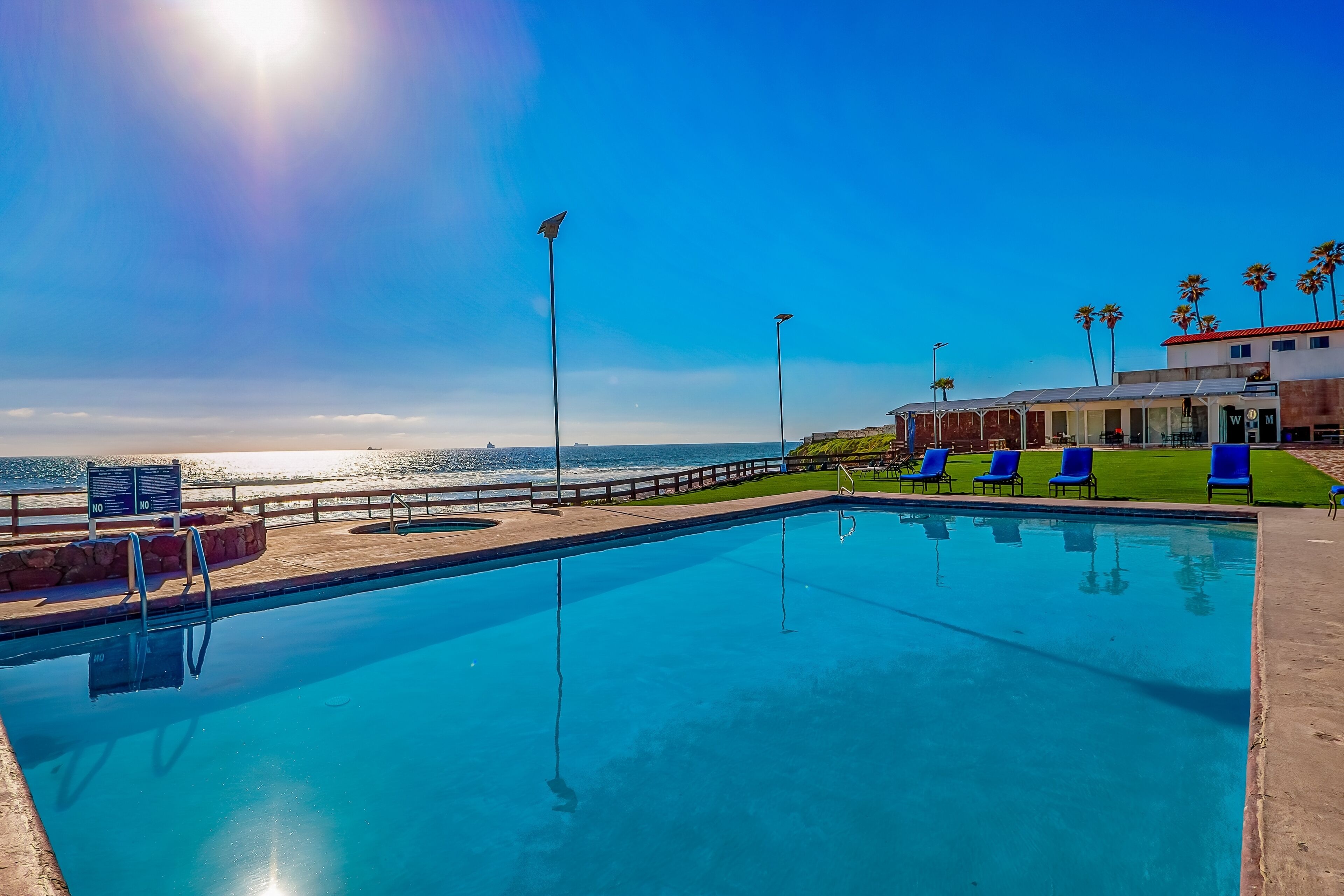 A swimming pool with a view of the ocean, blue lounge chairs, and a building with palm trees.