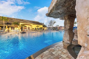 A swimming pool with a waterfall feature, a building with a tiled roof, and palm trees. - NEW! Minutes to Disney! Luxury Professionally Designed! Moana, Star Wars, Disney (Kissimmee)