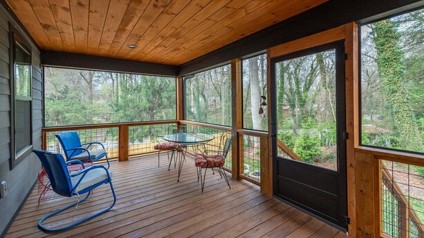 Terraza de madera con mesa y sillas, ventanas amplias y vista a los árboles.