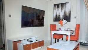 A modern room with a flat-screen TV, a dining table with orange chairs, a white coffee table, and a sofa. - Kindaruma Homes Apartments (Nairobi)