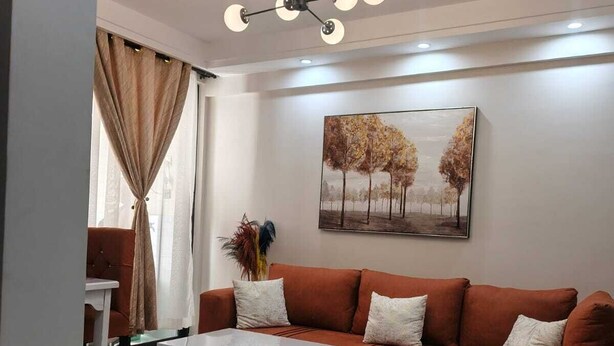 A living room with a sectional sofa, a coffee table, a painting on the wall, and a ceiling light fixture. - Kindaruma Homes Apartments (Nairobi)