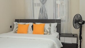 A bedroom with a bed, curtains, a nightstand, a fan, and a rug. - Kindaruma Homes Apartments (Nairobi)