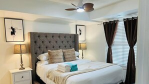 Un lit bien fait, doté d’une tête de lit capitonnée, de deux lampes de chevet, d’un ventilateur de plafond et d’une fenêtre avec des rideaux.