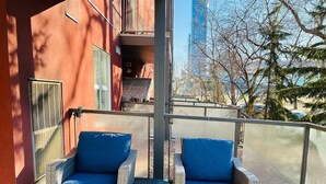 Un balcon agrémenté de fauteuils en osier et d’une petite table ronde, d’où l’on peut admirer un panorama urbain avec ses bâtiments et ses arbres.