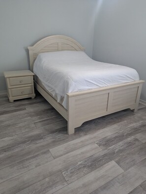 Un lit simple avec une tête de lit et un pied de lit, une table de chevet avec des tiroirs et un parquet à motifs chevrons gris.