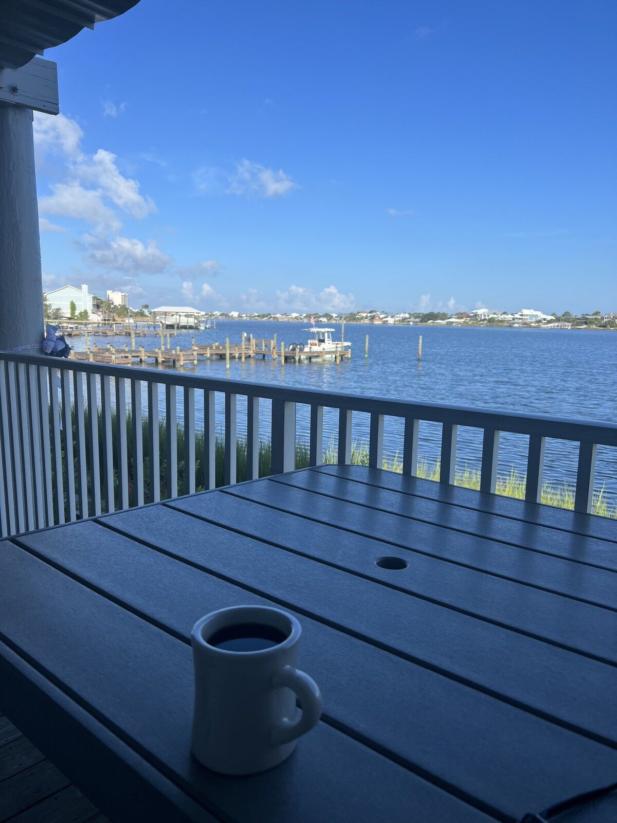 A wooden deck with a white mug, overlooking a calm body of water with docks and buildings in the distance.