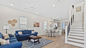 A living room with blue sofas, a coffee table, and a patterned rug. There is a staircase with white railings and a dining area with white furniture.