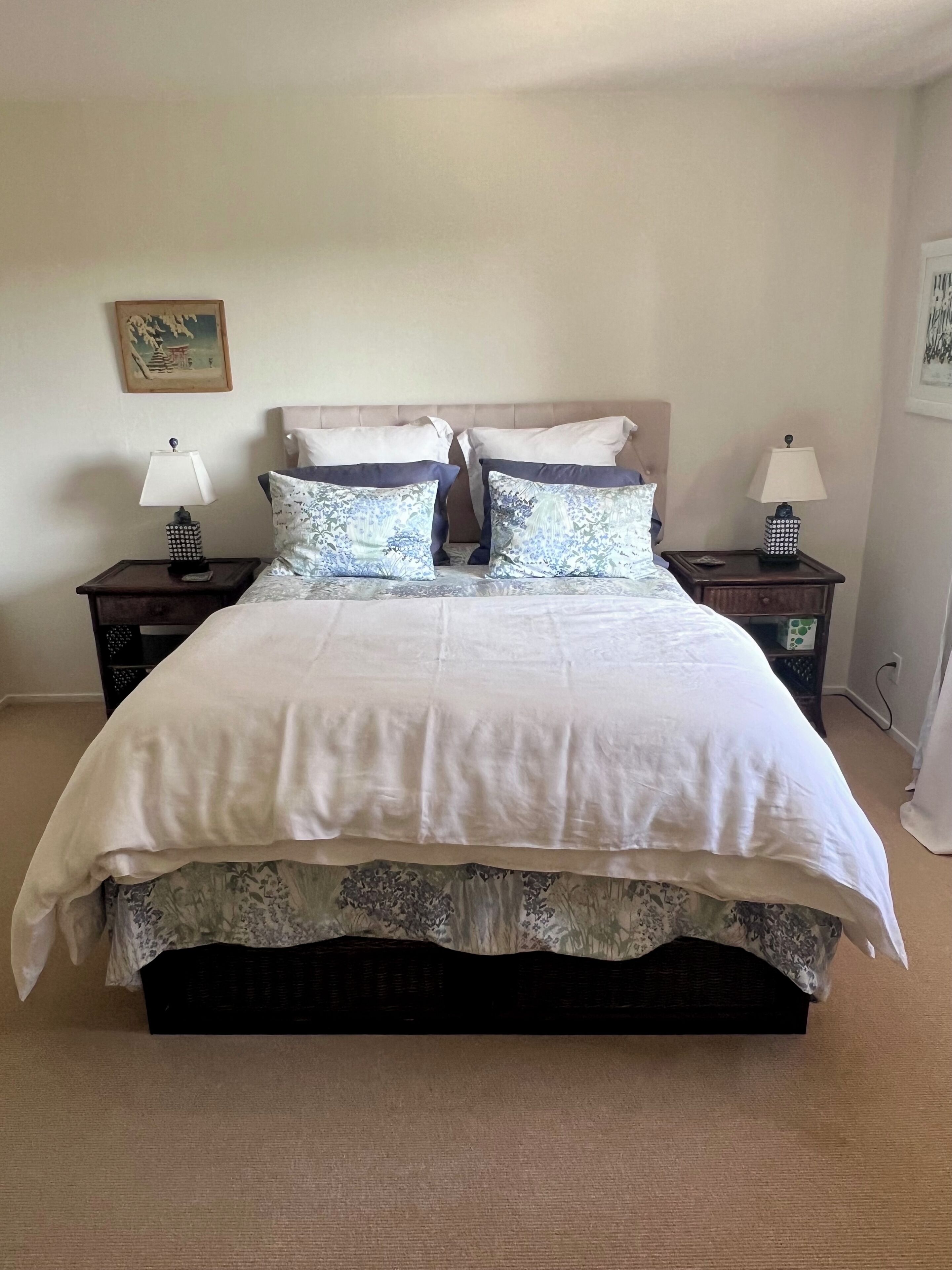 Una cama bien tendida con un edredón blanco y almohadas estampadas, flanqueada por dos mesitas de noche con lámparas, en una habitación con paredes lisas y suelo alfombrado.