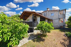 Une maison en pierre avec un porche en bois, un mur de pierre et un jardin agrémenté d’arbustes verts.