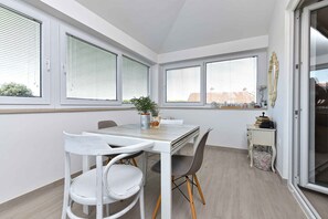 A dining area with a table and chairs, large windows with blinds, a small side table with books, and a plant on the table. - Holiday apartment tastefully furnished with air conditioning (Pula)