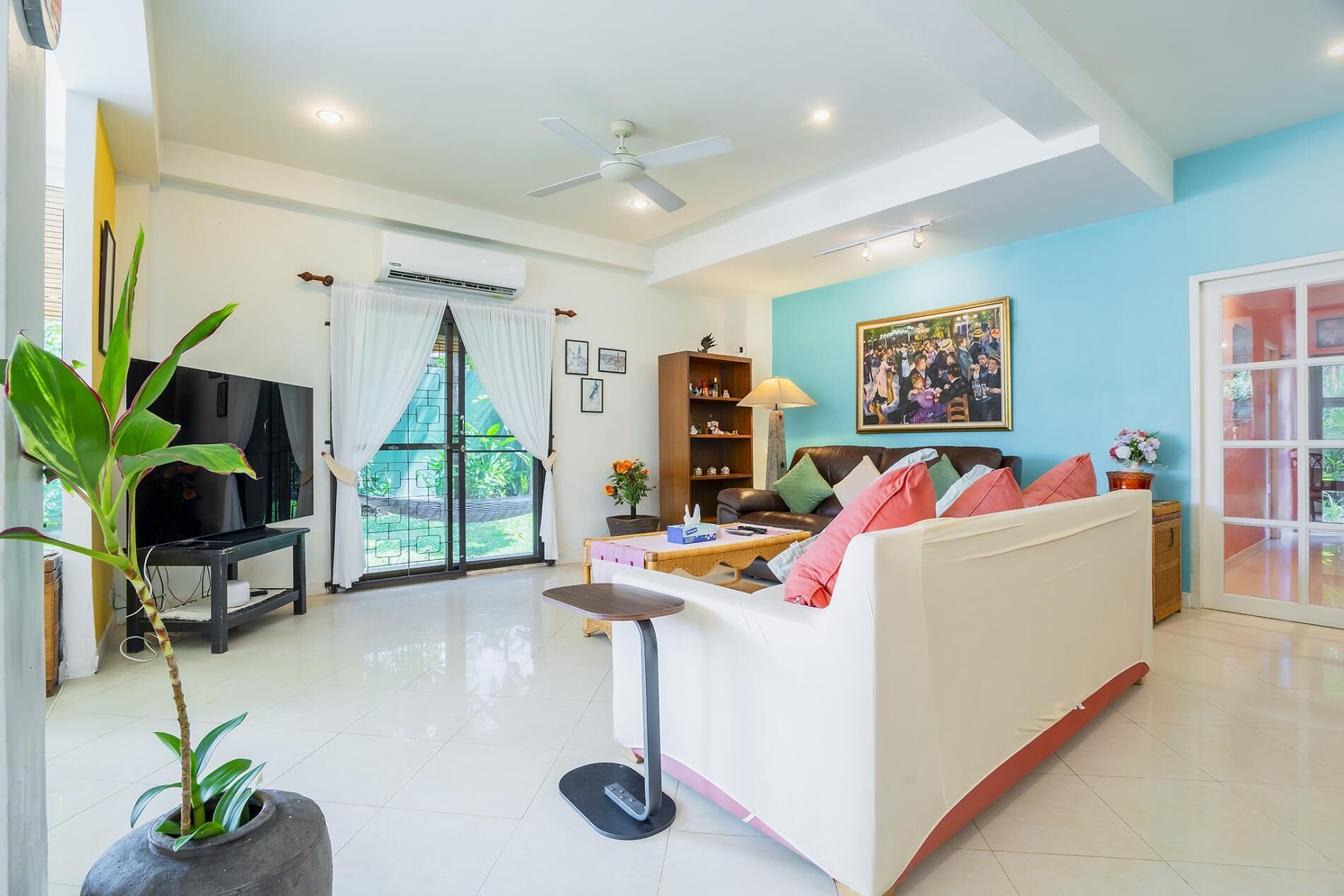 Un salon comprenant un canapé, un téléviseur, un ventilateur de plafond, une fenêtre avec des rideaux, une bibliothèque et un tableau accroché au mur.