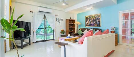 Un salon comprenant un canapé, un téléviseur, un ventilateur de plafond, une fenêtre avec des rideaux, une bibliothèque et un tableau accroché au mur.
