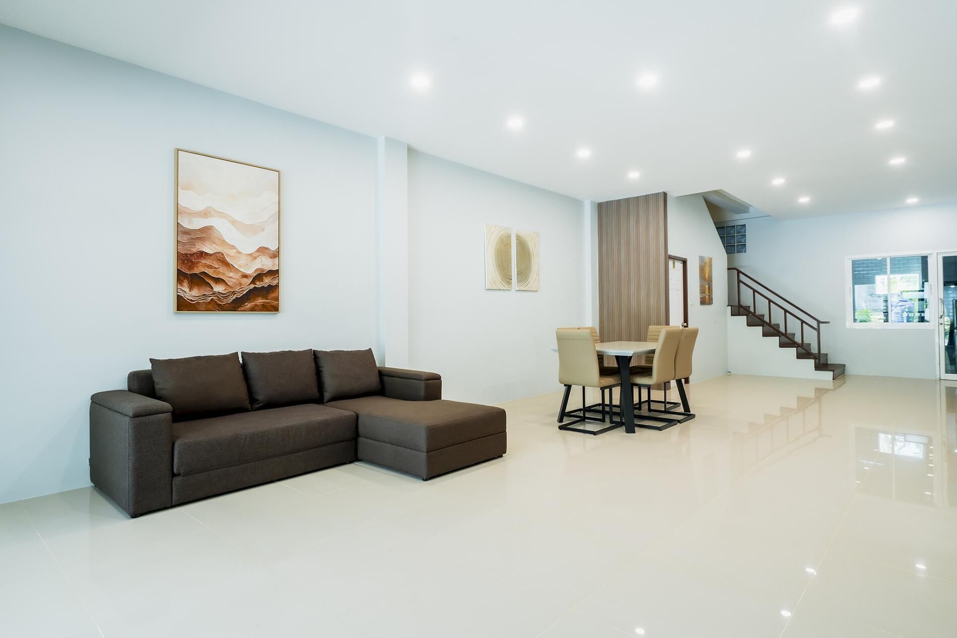 A modern living room with a sectional sofa, a dining table with chairs, a staircase, and artwork on the walls.