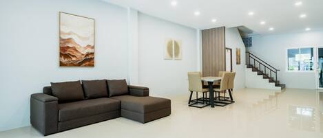A modern living room with a sectional sofa, a dining table with chairs, a staircase, and artwork on the walls.