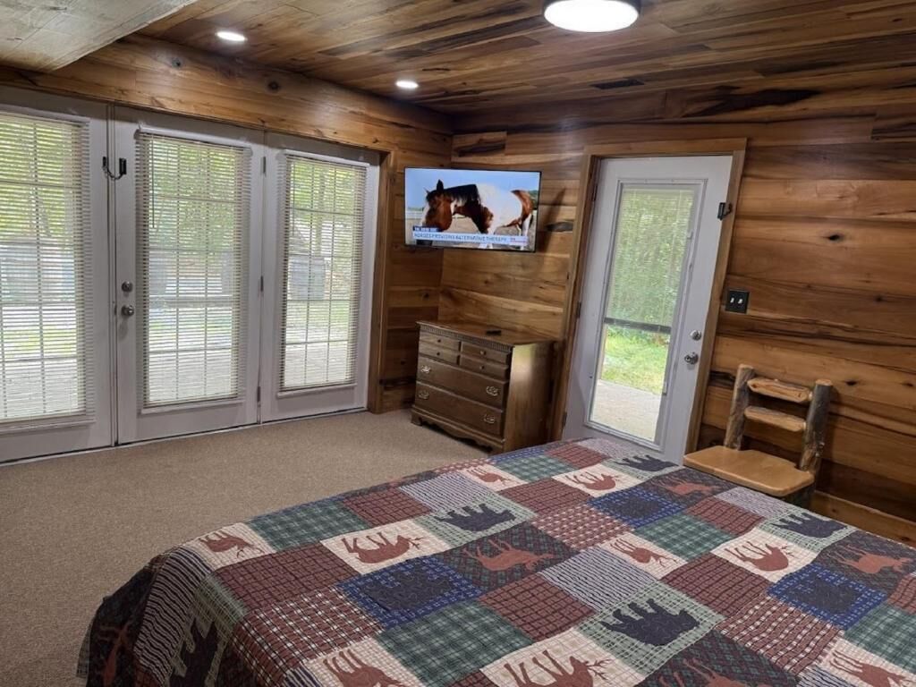 A bedroom with a wooden ceiling, a quilted bedspread, a television, a dresser, and a chair.