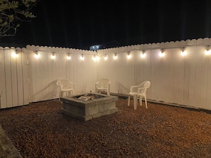 A patio with a fire pit and two chairs, surrounded by a white fence and string lights.